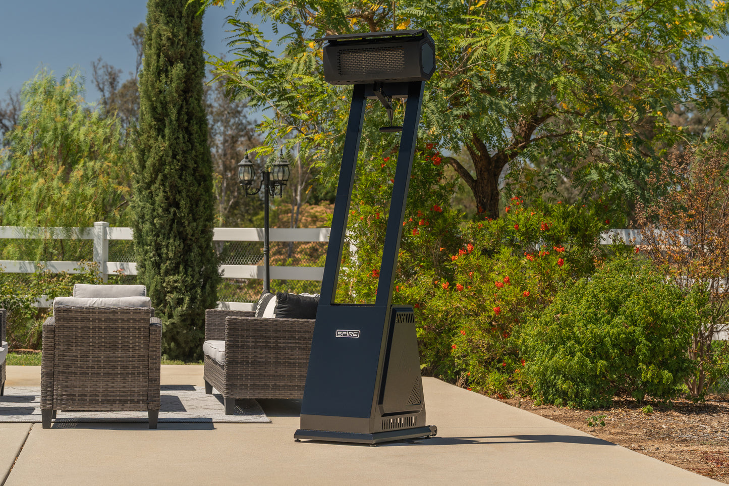 
                  
                    A freestanding outdoor patio heater with a dark navy blue body and a black top wind guard, positioned on a concrete patio in a landscaped backyard with wicker furniture and greenery.
                  
                